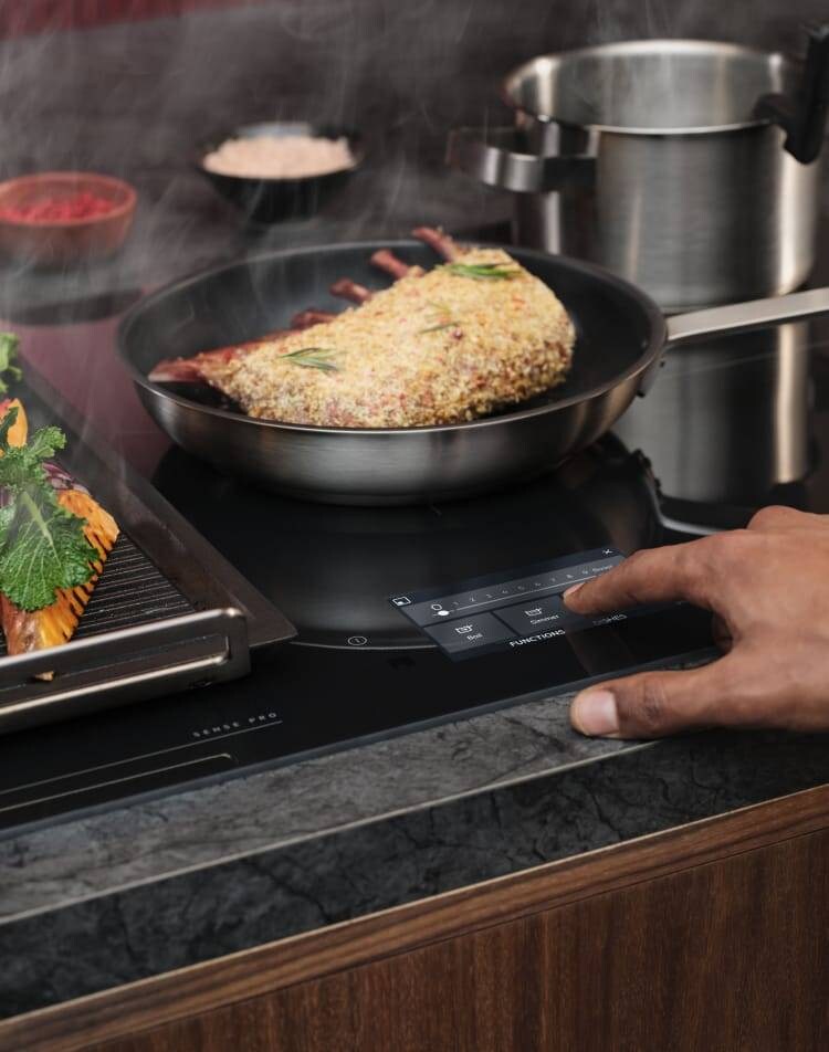 A finger is touching the control panel on an induction hob while food is being prepared.