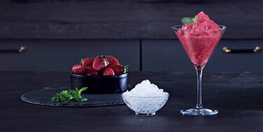 A strawberry daiquiri cocktail alongside a pile of strawberries and a bowl of ice